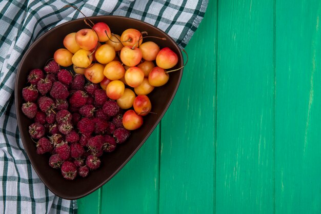 Vista superior de frambuesas con cerezas blancas en un recipiente con una toalla a cuadros sobre una superficie verde