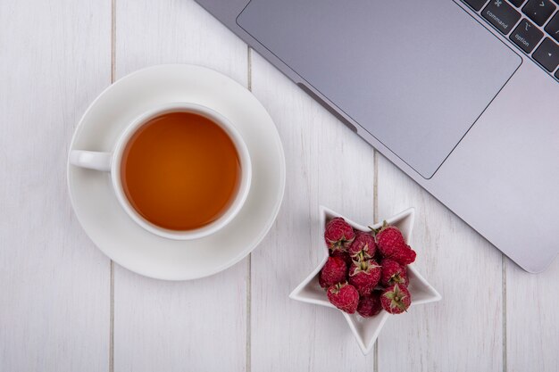 Vista superior de frambuesas con una taza de té y una computadora portátil sobre una superficie blanca