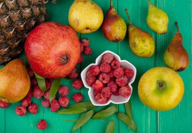 Vista superior de frambuesas en un tazón con granada, piña, durazno y manzana con hojas alrededor en la superficie verde