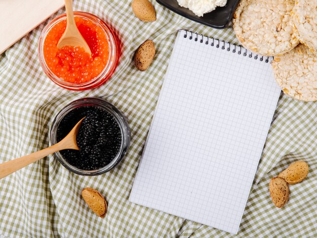 Vista superior frasco de caviar rojo y negro con cucharas de madera crujiente de almendras requesón y espacio de copia en la mesa