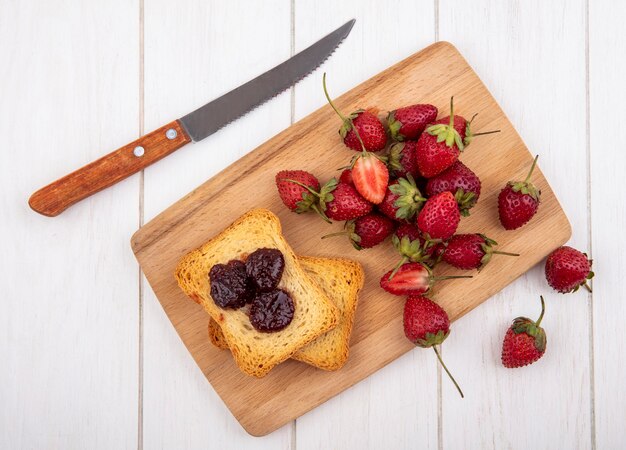 Vista superior de fresa fresca con pan tostado en una tabla de cocina de madera con cuchillo sobre un fondo blanco.