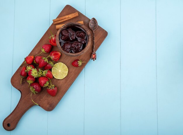 Vista superior de fresa fresca en una tabla de cocina de madera con mermelada de fresa con una rodaja de limón sobre un fondo azul con espacio de copia