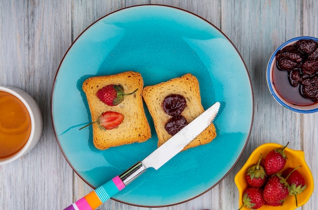 Vista superior de la fresa en el pan en un plato azul con un cuchillo con mermelada de fresa en un recipiente con fresas frescas sobre un fondo de madera gris