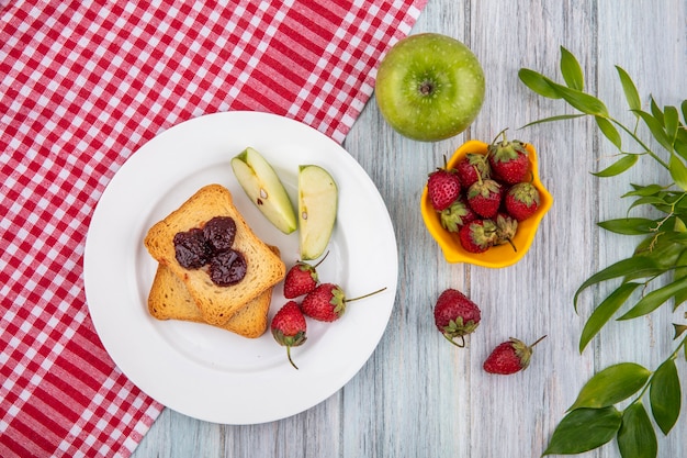 Vista superior de la fresa en un plato blanco con manzanas verdes sobre un mantel de cuadros rojos con fresas frescas en un recipiente amarillo con hojas sobre un fondo de madera gris