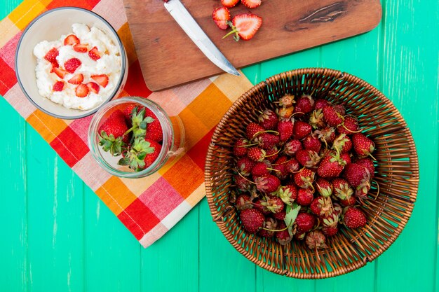 Vista superior de fresas en la cesta con cuencos de requesón y fresas en tela sobre superficie verde