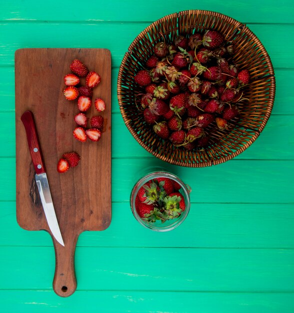 Vista superior de fresas cortadas con un cuchillo en la tabla de cortar y fresas enteras en la cesta y el tazón sobre una superficie verde