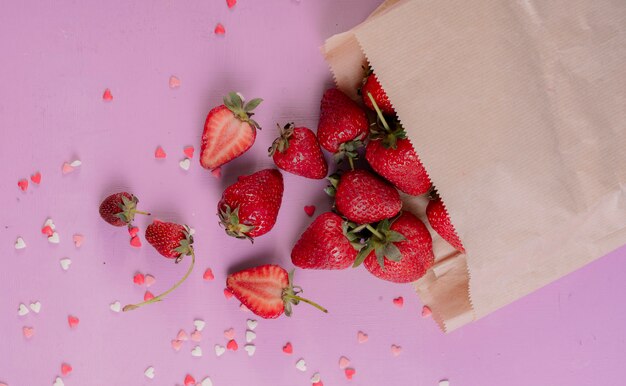 Vista superior de fresas cortadas y enteras derramándose de una bolsa de papel sobre una mesa morada