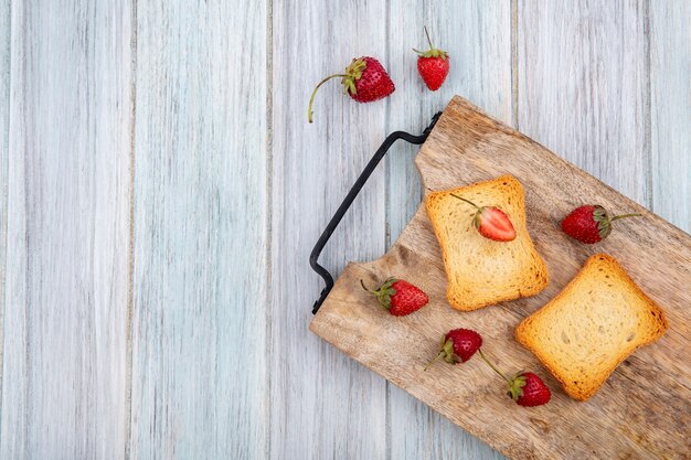 Vista superior de fresas frescas y deliciosas con rebanadas de pan tostado en una tabla de cocina de madera sobre un fondo de madera gris con espacio de copia