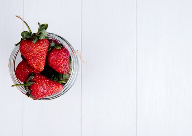 Vista superior de fresas frescas maduras en un frasco de vidrio sobre una superficie blanca con espacio de copia