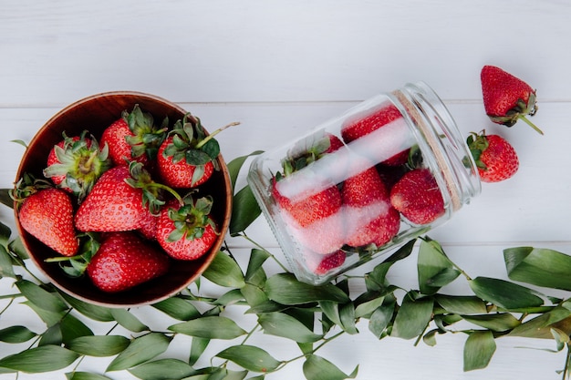 Vista superior de fresas frescas maduras en un tazón de madera y fresas esparcidas de un frasco de vidrio y hojas verdes en la mesa de madera blanca