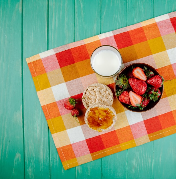 Vista superior de fresas frescas maduras en un tazón de madera y pasteles de arroz con mermelada y un vaso de leche sobre tela a cuadros en madera verde con espacio de copia