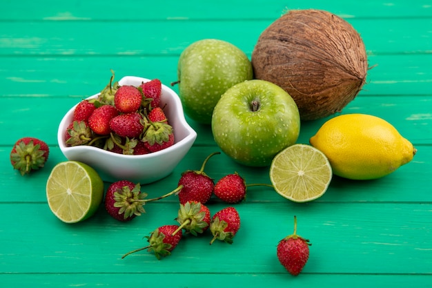 Vista superior de fresas frescas en un recipiente blanco con manzanas verdes, coco, limón, lima sobre un fondo de madera verde