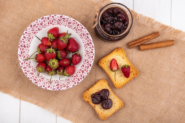 Vista superior de fresas frescas en un recipiente sobre un paño de saco con mermelada de fresa con ramas de canela sobre un fondo de madera blanca