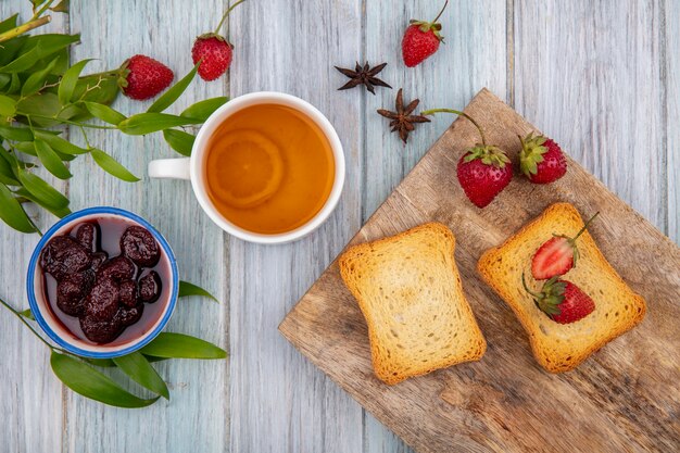 Vista superior de fresas frescas en una tabla de cocina de madera con rebanadas de pan tostado con mermelada de fresa con una taza de té sobre un fondo gris de madera