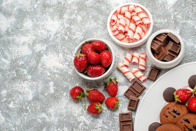 Vista superior fresas galletas y bombones redondos en el plato blanco y cuencos con bombones de fresas dulces en el suelo gris-blanco