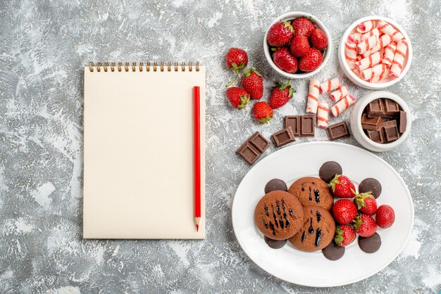 Vista superior fresas galletas y bombones redondos en el plato blanco y cuencos de dulces fresas chocolates y cuaderno sobre el suelo gris-blanco