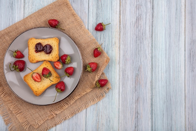 Vista superior de fresas en pan tostado en un plato con fresas frescas en un paño de saco sobre un fondo de madera gris con espacio de copia