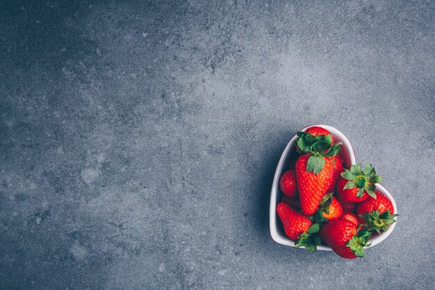 Vista superior de fresas en un recipiente en forma de corazón sobre fondo gris con textura. espacio libre horizontal para su texto