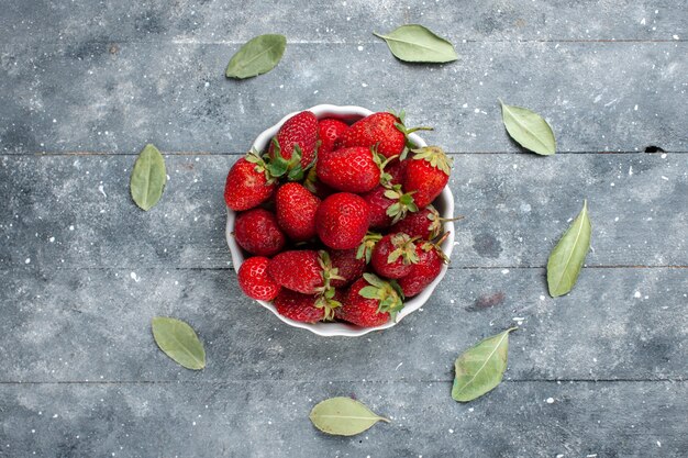 Vista superior de fresas rojas frescas dentro de la placa blanca junto con hojas secas verdes en madera gris, salud de vitamina baya fresca de frutas