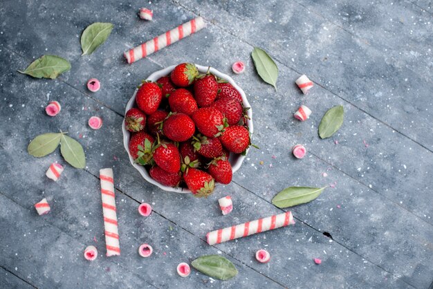 Vista superior de fresas rojas frescas dentro de la placa junto con caramelos de palo en gris, dulce de caramelo fresco de baya de fruta