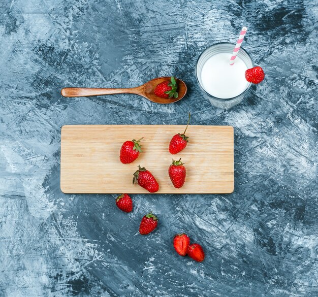 Vista superior de fresas en tabla de cortar con leche y una cuchara de madera sobre superficie de mármol azul oscuro. horizontal