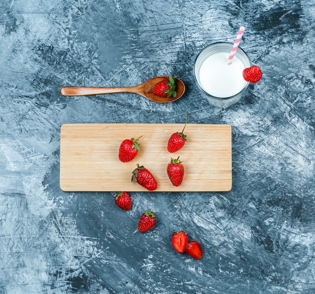 Vista superior de fresas en tabla de cortar con leche y una cuchara de madera sobre superficie de mármol azul oscuro. horizontal