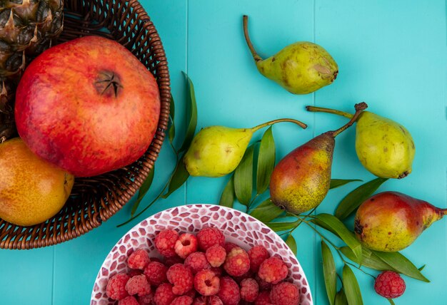 Vista superior de frutas en canasta y tazón de frambuesa con duraznos y hojas sobre superficie azul