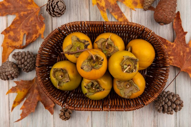 Vista superior de frutas de caqui en un balde con hojas aisladas sobre una mesa de madera gris