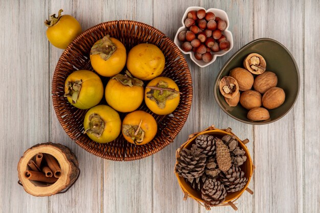 Vista superior de frutas de caqui en un balde con piñas en un balde con avellanas en un recipiente con ramas de canela sobre una mesa de madera gris