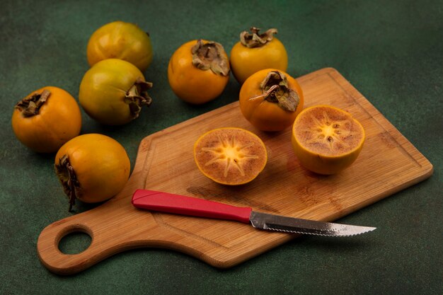 Vista superior de las frutas de caqui a la mitad en una tabla de cocina de madera con un cuchillo con frutas de caqui enteras aisladas sobre una superficie verde