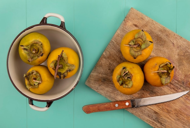 Vista superior de frutas de caqui naranja en una tabla de cocina de madera con cuchillo con frutas de caqui en un recipiente sobre una mesa de madera azul