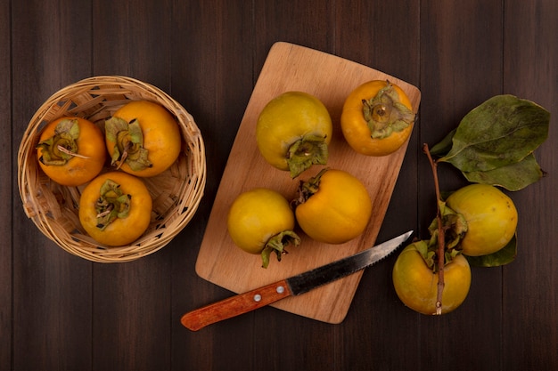 Vista superior de frutas de caqui orgánico en un balde con frutas de caqui en una tabla de cocina de madera con cuchillo sobre una mesa de madera