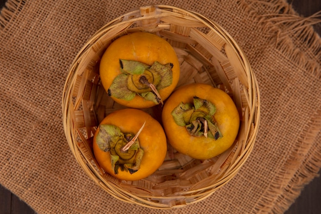 Foto gratuita vista superior de frutas de caqui orgánico en un balde sobre un paño de saco sobre una mesa de madera