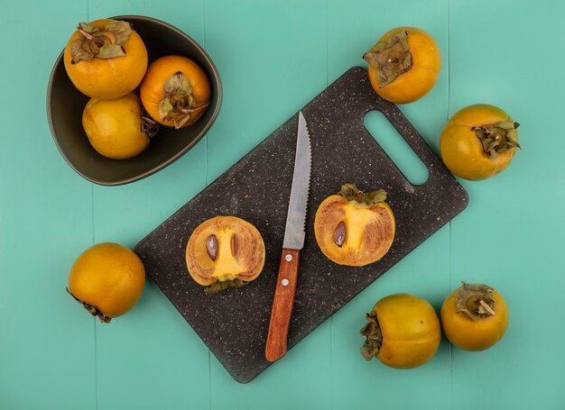 Vista superior de frutas de caqui redondeado naranja sobre una placa de cocina negra con cuchillo con frutas de caqui en un recipiente sobre una mesa de madera azul