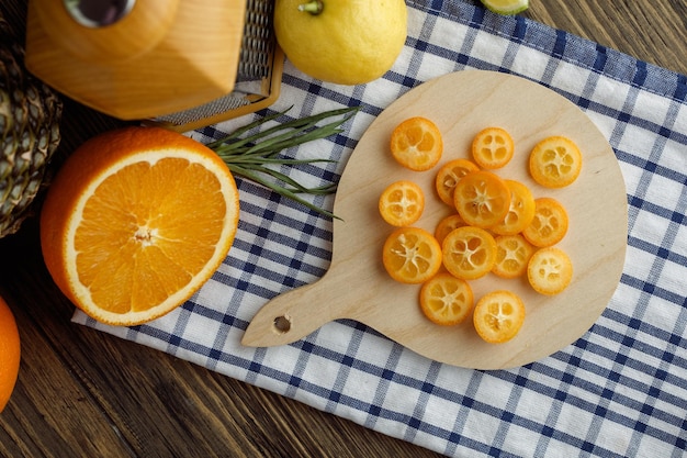 Foto gratuita vista superior de frutas cítricas como rodajas de kumquat en tabla de cortar y cebolleta de naranja cortada con limón con rallador sobre tela escocesa sobre fondo de madera