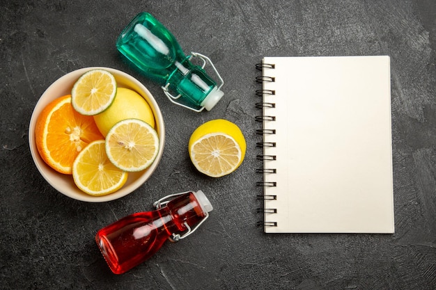 Vista superior de frutas cítricas frutas cítricas en un tazón junto al cuaderno blanco y dos botellas en la mesa oscura