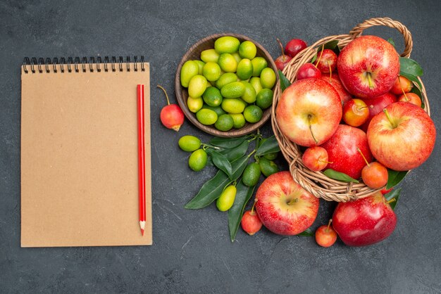 Vista superior frutas cítricos cesta de madera de cerezas manzanas crema cuaderno lápiz rojo