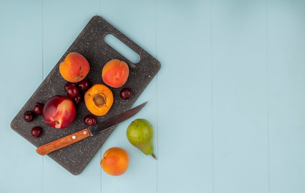 Vista superior de frutas como albaricoques enteros y la mitad y cerezas de durazno con un cuchillo en la tabla de cortar y albaricoque de pera sobre fondo azul con espacio de copia