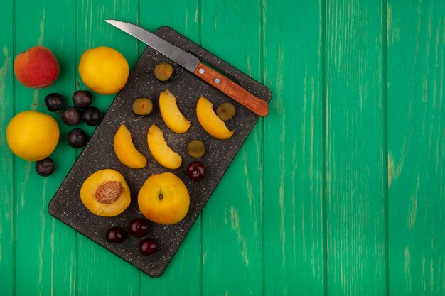 Vista superior de las frutas como albaricoques en rodajas medias enteras y endrinas con un cuchillo sobre la tabla de cortar y sobre fondo verde con espacio de copia