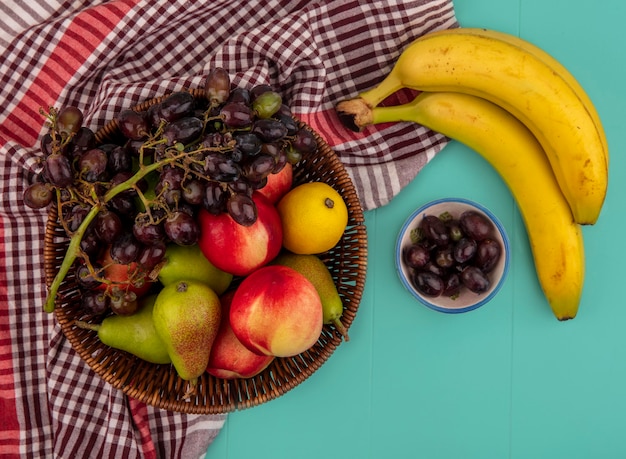 Foto gratuita vista superior de frutas como canasta de uva pera melocotón manzana limón sobre tela escocesa con plátanos y bayas de uva sobre fondo azul.