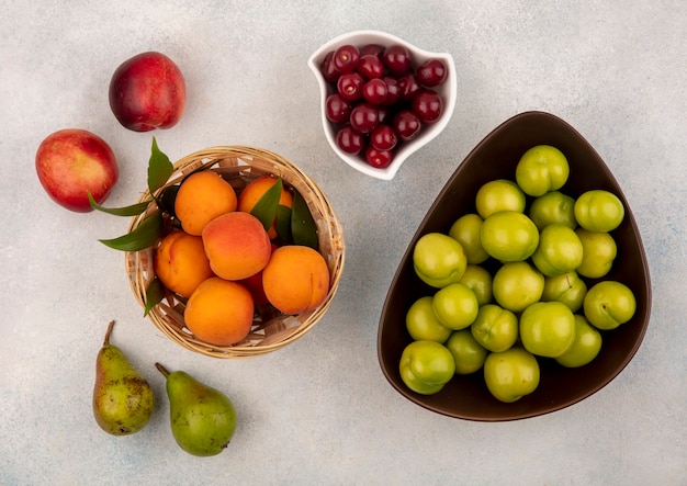 Vista superior de frutas como cereza de albaricoque y ciruela en canasta y cuencos con melocotones y peras sobre fondo blanco.