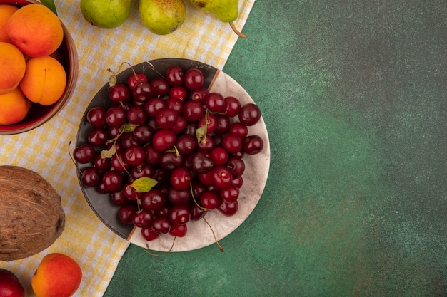 Vista superior de frutas como cerezas y albaricoques en plato y tazón con peras y coco en tela escocesa sobre fondo verde con espacio de copia