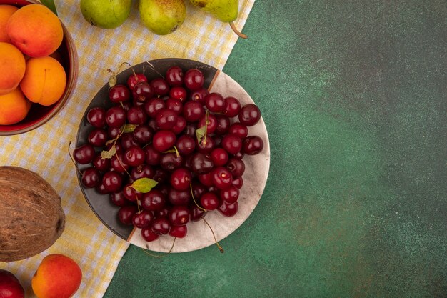 Vista superior de frutas como cerezas y albaricoques en plato y tazón con peras y coco en tela escocesa sobre fondo verde con espacio de copia