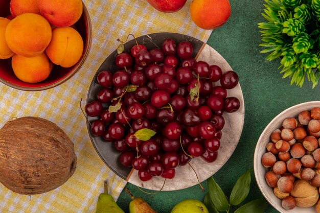 Vista superior de frutas como cerezas y albaricoques en plato y tazón con peras y coco en tela escocesa y tazón de nueces sobre fondo verde