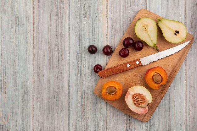 Vista superior de frutas como cerezas y medio corte albaricoque pera y melocotón con cuchillo sobre tabla de cortar sobre fondo de madera con espacio de copia