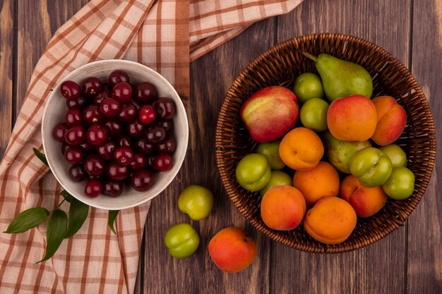 Vista superior de frutas como cerezas en un tazón sobre tela escocesa y canasta de melocotón, albaricoque, pera, ciruela sobre fondo de madera