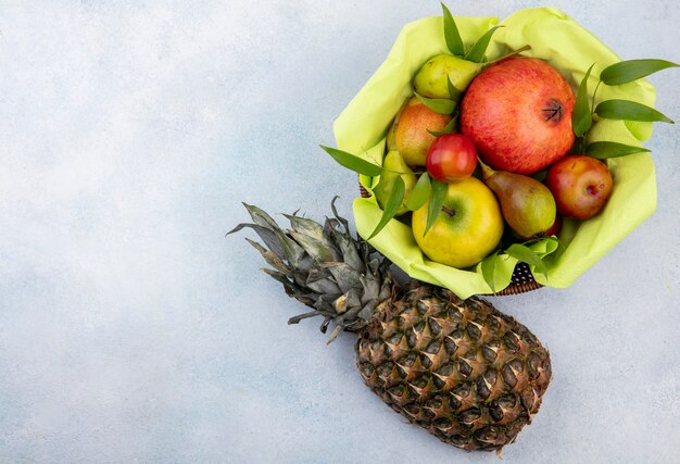 Vista superior de frutas como ciruela manzana durazno y granada en canasta con piña en superficie blanca con espacio de copia