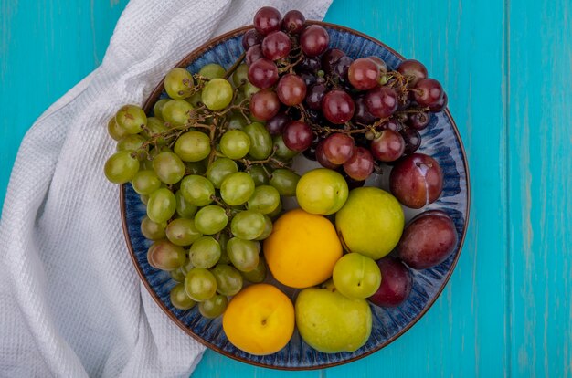 Vista superior de frutas como ciruelas y uvas pluots nectacots en placa sobre tela blanca sobre fondo azul.