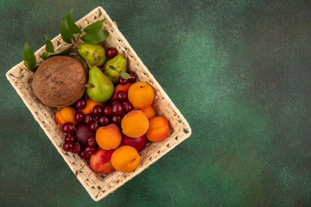 Vista superior de frutas como coco melocotón albaricoque pera cereza con hojas en canasta sobre fondo verde con espacio de copia