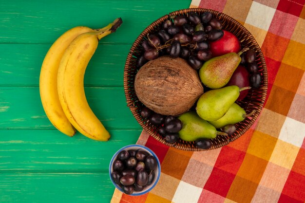 Vista superior de frutas como coco pera uva melocotón en canasta sobre tela escocesa y plátanos sobre fondo verde